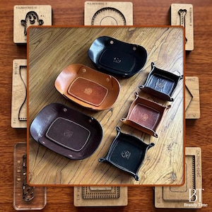May include: A collection of leather valet trays in various shapes and colors, including black, tan, and dark brown. The trays feature metal snap fasteners at the corners and a circular embossed logo in the center of each base, arranged on a surface.