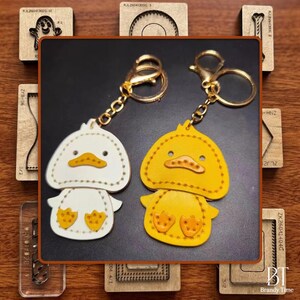 Könnte beinhalten: Zwei Leder-Enten-Schlüsselanhänger, einer weiß und einer gelb, mit goldfarbener Hardware. Die Enten haben gestickte Details und befinden sich auf einer schwarzen Oberfläche. Holzhandwerkzeuge umgeben die Schlüsselanhänger.