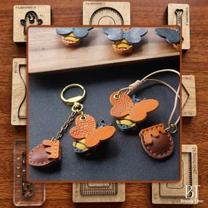 May include: Handmade leather bee-themed accessories, including keychains and charms. The bees have orange wings and yellow and black bodies. A small leather honey pot charm is also shown. The background includes wooden craft tools.