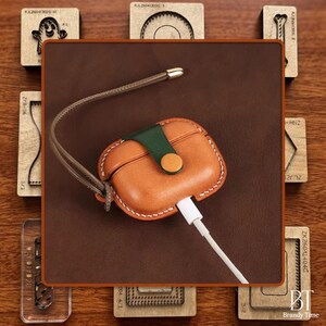 Könnte beinhalten: Eine braune Leder-AirPod-Hülle mit grünem Riemen und Knopfverschluss. Die Hülle hat weiße Nähte und einen geflochtenen Riemen. Ein weißes Ladekabel ist eingesteckt. Der Hintergrund umfasst Holzblöcke.
