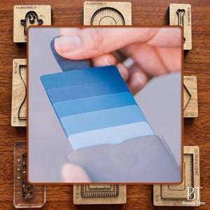May include: A hand holding a fan of blue color swatches, ranging from light to dark shades. The swatches are displayed against a neutral background. Various wooden craft stamps are arranged around the swatches.
