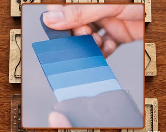 May include: A hand holding a fan of blue color swatches, ranging from light to dark shades. The swatches are displayed against a neutral background. Various wooden craft stamps are arranged around the swatches.