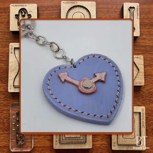 May include: A heart-shaped leather keychain in light blue with a pink clock design. The keychain has a silver chain and is surrounded by wooden craft tools. The design features stitched edges.