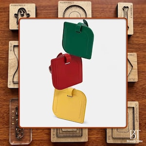 Peut inclure: Trois étiquettes de bagage en cuir vert, rouge et jaune sont empilées sur un fond blanc. Chaque étiquette a une forme rectangulaire avec un dessus incurvé et une petite sangle. Les étiquettes sont entourées d'outils artisanaux en bois.