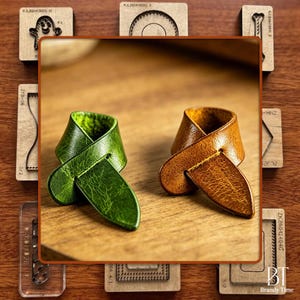 May include: Two leather guitar straps, one green and one brown, are displayed on a wooden surface. The green strap has a textured appearance, while the brown strap has a smooth finish. The straps are next to wooden stamps.