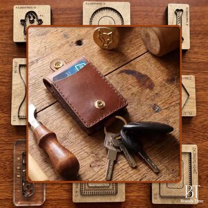 May include: A brown leather cardholder with a gold snap closure, holding a credit card, next to a set of keys. The cardholder is on a wooden surface, surrounded by leatherworking tools and stamps.