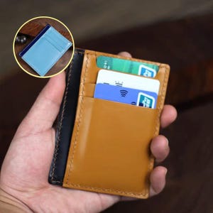 Puede incluir: Un tarjetero de cuero marrón claro con múltiples ranuras para tarjetas, que contiene varias tarjetas de crédito. El tarjetero tiene un lomo negro y un interior azul claro. Una pequeña incrustación circular muestra el diseño interior.