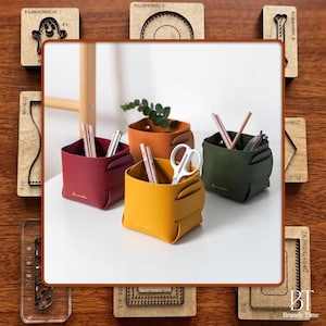 Puede incluir: Cuatro cubos de almacenamiento cuadrados y coloridos en rojo, naranja, amarillo y verde, cada uno con un asa pequeña. Los cubos están llenos de lápices, tijeras y pequeñas plantas. También se ven sellos de madera.