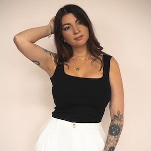 Könnte beinhalten: Ein schwarzes Tanktop mit quadratischem Ausschnitt und eine weiße Hose mit hohem Bund. Das Top ist ärmellos und figurbetont. Die Hose hat ein weites Bein und einen Knopfverschluss. Das Outfit ist schlicht und stilvoll.