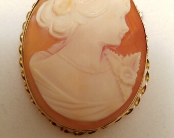 May include: Oval cameo brooch featuring a profile of a woman with detailed hair and clothing. The cameo is set in a gold-colored twisted rope frame. The woman's profile is carved in a light peach color, set against a darker peach background.