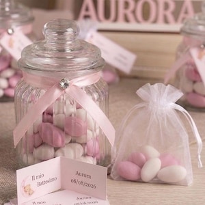 Può includere: Barattoli di vetro pieni di caramelle rosa e bianche, legati con nastri rosa e un accento di strass. I barattoli fanno parte di una celebrazione battesimale, con il nome "Aurora" esposto sullo sfondo. Sono presenti anche piccole carte con testo.