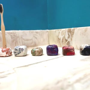 May include: A bamboo toothbrush with black bristles stands upright in a small, round, marbled clay holder. Beside it are five additional clay holders in various marbled color combinations, including white, gray, green, brown, purple, and black.