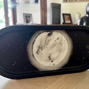 Puede incluir: Una bandeja ovalada de color negro que presenta una incrustación circular central con un patrón jaspeado en blanco y gris. La superficie negra de la bandeja está salpicada de pequeños puntos blancos, creando contraste con el círculo central claro.