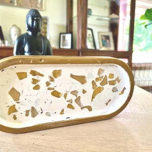 Könnte beinhalten: Ein ovales Tablett mit einem goldfarben lackierten Rand und einer weißen Innenfläche. Die Innenfläche ist mit verstreuten, unregelmäßigen Stücken aus bernsteinfarbenem Glas durchsetzt, was einen mosaikartigen Effekt im weißen Material erzeugt.