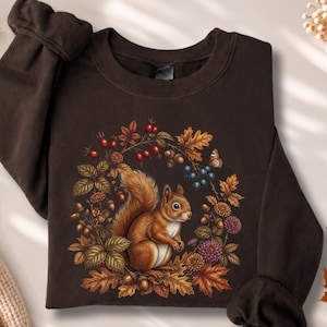 May include: A dark brown crewneck sweatshirt featuring a central graphic of a brown squirrel surrounded by a wreath of autumn leaves, red berries, blue berries, acorns, and purple flowers. The long-sleeved garment has ribbed cuffs and a ribbed neckline.