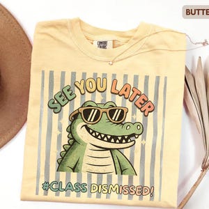 May include: Butter-colored t-shirt featuring a cartoon alligator wearing sunglasses. The shirt has the text "SEE YOU LATER" and "#CLASS DISMISSED!". The design is set against a striped background.
