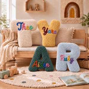 May include: Four plush letter-shaped pillows in cream, yellow, dark green, and light blue. The cream T features the name Theo in blue and green, the yellow Y features Yara in purple and orange, the green A features Aiden in orange and pink, and the blue R features Ruby in pink and purple.