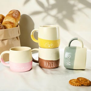 Może przedstawiać: Cztery ceramiczne kubki w różnych kolorach, w tym żółtym, różowym, brązowym i zielonym, z nadrukowanymi imionami. Kubki są ułożone jeden na drugim i ustawione na białej powierzchni, z papierową torbą z chlebem i ciastkami w tle.