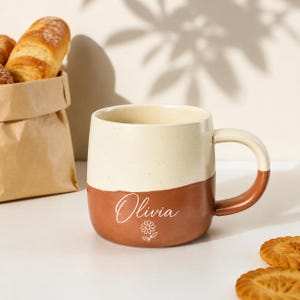 Könnte beinhalten: Ein zweifarbiger Keramikbecher mit einem gesprenkelten cremefarbenen oberen Teil und einem terrakottafarbenen unteren Teil. Auf der Vorderseite steht der Name Olivia in weißer Schreibschrift über einer kleinen, weißen Gänseblümchen-Grafik.