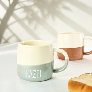 Puede incluir: Tazas de cerámica de dos tonos con asas. Una taza es crema y verde azulado con el nombre "HAZEL" en blanco. La otra taza es crema y marrón con un nombre en cursiva. Las tazas están sobre una superficie blanca.