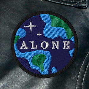 May include: Embroidered patch featuring a blue and green globe design with the word "ALONE" in white. The patch includes two white star-like shapes and is bordered by a black trim. The patch is circular and appears to be attached to a dark leather jacket.