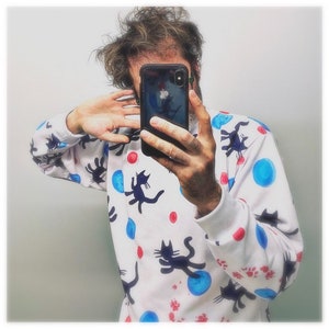 Puede incluir: Una sudadera blanca con estampado de gato negro y lunares azules y rojos. La sudadera tiene cuello redondo y mangas largas.