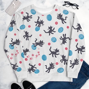 Könnte beinhalten: Weißer Pullover mit einem schwarzen Katzen- und blau-roten Kreis-Muster.