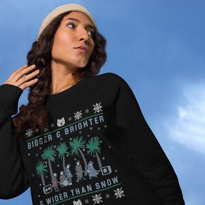 Op de afbeelding: Een zwart sweatshirt met een wit, groen en blauw kersttrui-ontwerp. Het ontwerp bevat palmbomen, een band van mensen die instrumenten bespelen, en de tekst "Bigger & Brighter & Wider Than Snow".