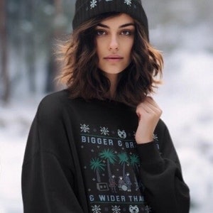 Op de afbeelding: Een zwarte beanie met een pompon en een bijpassende zwarte sweatshirt. De beanie heeft een beren- en sneeuwvlokkenpatroon. De sweatshirt heeft een afbeelding met de tekst "BIGGER & BR" en "& WIDER THAN". De afbeelding toont palmbomen en een band.