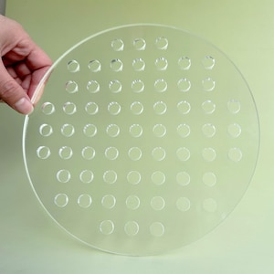 Puede incluir: Una lámina circular transparente de acrílico con numerosos agujeros espaciados uniformemente. El material transparente permite el paso de la luz, creando un patrón de rejilla. El objeto es sostenido por una mano, mostrando su tamaño y forma.