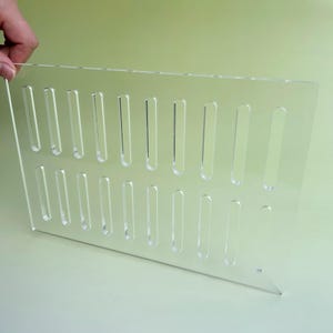 Può includere: Un espositore in acrilico trasparente con numerose fessure verticali. Il supporto è tenuto da una mano ed è progettato per esporre oggetti. Il materiale trasparente offre una visione chiara dei prodotti esposti.