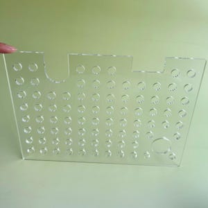 Puede incluir: Organizador de acrílico transparente con múltiples agujeros circulares. Tiene forma rectangular con un recorte curvo en un lado y una parte superior plana con un pequeño recorte. El material transparente permite la visibilidad del contenido.