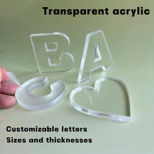 May include: Clear acrylic letters and a heart shape are displayed against a light green background. The letters spell out "BA" and the text "Transparent acrylic", "Customizable letters", and "Sizes and thicknesses" are also visible.