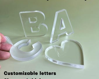 May include: Transparent acrylic letters and a heart shape are shown on a pale green surface. The letters spell "BA". Text reads "Transparent acrylic", "Customizable letters", and "Sizes and thicknesses".