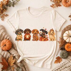 May include: A cream-colored t-shirt featuring a graphic of five dachshunds. The dogs are depicted wearing autumn-themed accessories including a pumpkin-patterned sweater, a red sweater with a mug, a plaid scarf, playing in leaves, and an acorn hat.