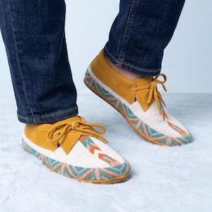 May include: Mustard yellow moccasins featuring a white beaded vamp and toe box with a geometric pattern in teal and orange. The shoes have matching yellow laces tied in a bow at the front and a low-profile silhouette with a flat sole.