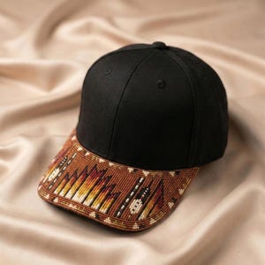 May include: A black baseball cap featuring a structured crown and a curved brim adorned with intricate, multi-coloured beadwork. The bead pattern displays a geometric design in shades of brown, orange, yellow, and white, covering the entire brim.
