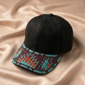 Puede incluir: Una gorra de béisbol negra con una corona estructurada y una visera curva. La visera está decorada con un intrincado patrón de pequeñas cuentas en tonos turquesa, naranja y granate oscuro, dispuestas en un diseño geométrico sobre la superficie.