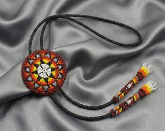 Puede incluir: Una corbata bolo con un colgante circular de cuentas rojas, naranjas, amarillas y blancas, con un diseño de sol. El colgante está unido a un cordón trenzado negro con puntas de cuentas.