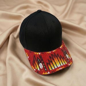 May include: A black baseball cap featuring a curved brim adorned with intricate beadwork in a vibrant pattern of red, orange, yellow, and white. The crown of the cap is solid black fabric with a button at the top and stitched eyelets for ventilation.