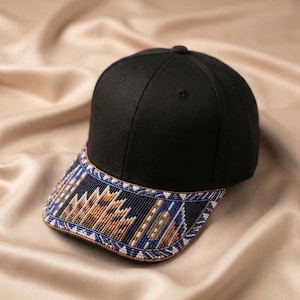 Puede incluir: Una gorra de béisbol negra con una visera curva adornada con intrincados abalorios en tonos azul, beige y blanco. El trabajo de cuentas muestra un patrón geométrico que cubre toda la superficie de la visera, creando una apariencia texturizada.