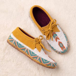 May include: A pair of mustard yellow suede moccasins featuring white, teal, and orange beadwork in a geometric pattern. The shoes have a lace-up front with matching yellow suede ties and a dark purple interior lining with a flat sole.