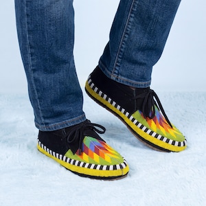 May include: Black suede moccasins featuring a vibrant, multi-colored beaded geometric pattern on the toe and a black and white striped beaded border along the sole. The shoes include black laces and are worn with blue denim jeans.
