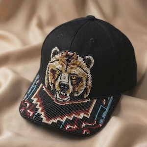Puede incluir: Una gorra de béisbol negra con un diseño detallado de cabeza de oso en el panel frontal, elaborado con pequeñas cuentas en tonos marrón, dorado y blanco. La visera está decorada con un patrón geométrico de cuentas en negro, rojo, blanco y azul.