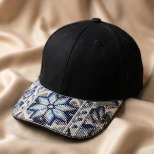 Puede incluir: Una gorra de béisbol negra con una visera curva adornada con un detallado patrón de cuentas multicolor. El trabajo de abalorios muestra un diseño geométrico similar a un copo de nieve en tonos azules, blancos y grises sobre la visera.