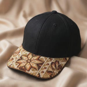 May include: A black baseball cap featuring a structured crown and a curved brim adorned with intricate beadwork in shades of tan, brown, and cream, arranged in a geometric star-like pattern that covers the entire surface of the brim.