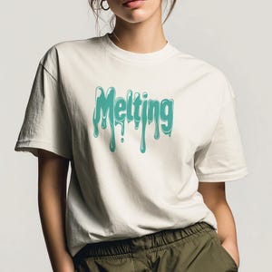May include: Off-white t-shirt with the word "Melting" in turquoise, dripping font. The short-sleeved shirt is made of a soft material and is worn by a person. The person is wearing olive green pants.