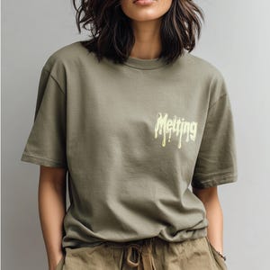 May include: Olive green t-shirt with the word "Melting" in a dripping yellow font on the upper chest. The shirt has short sleeves and a relaxed fit. The model is wearing olive green pants.