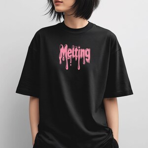 May include: Black t-shirt with the word "Melting" in pink, dripping font across the chest. The shirt has short sleeves and a crew neck. The model is wearing the shirt.
