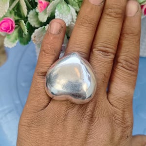 Puede incluir: Un anillo grande plateado en forma de corazón. El anillo tiene una superficie lisa y pulida que refleja la luz. El anillo se lleva en un dedo. El fondo incluye una superficie azul y un arreglo floral con flores rosas y blancas.